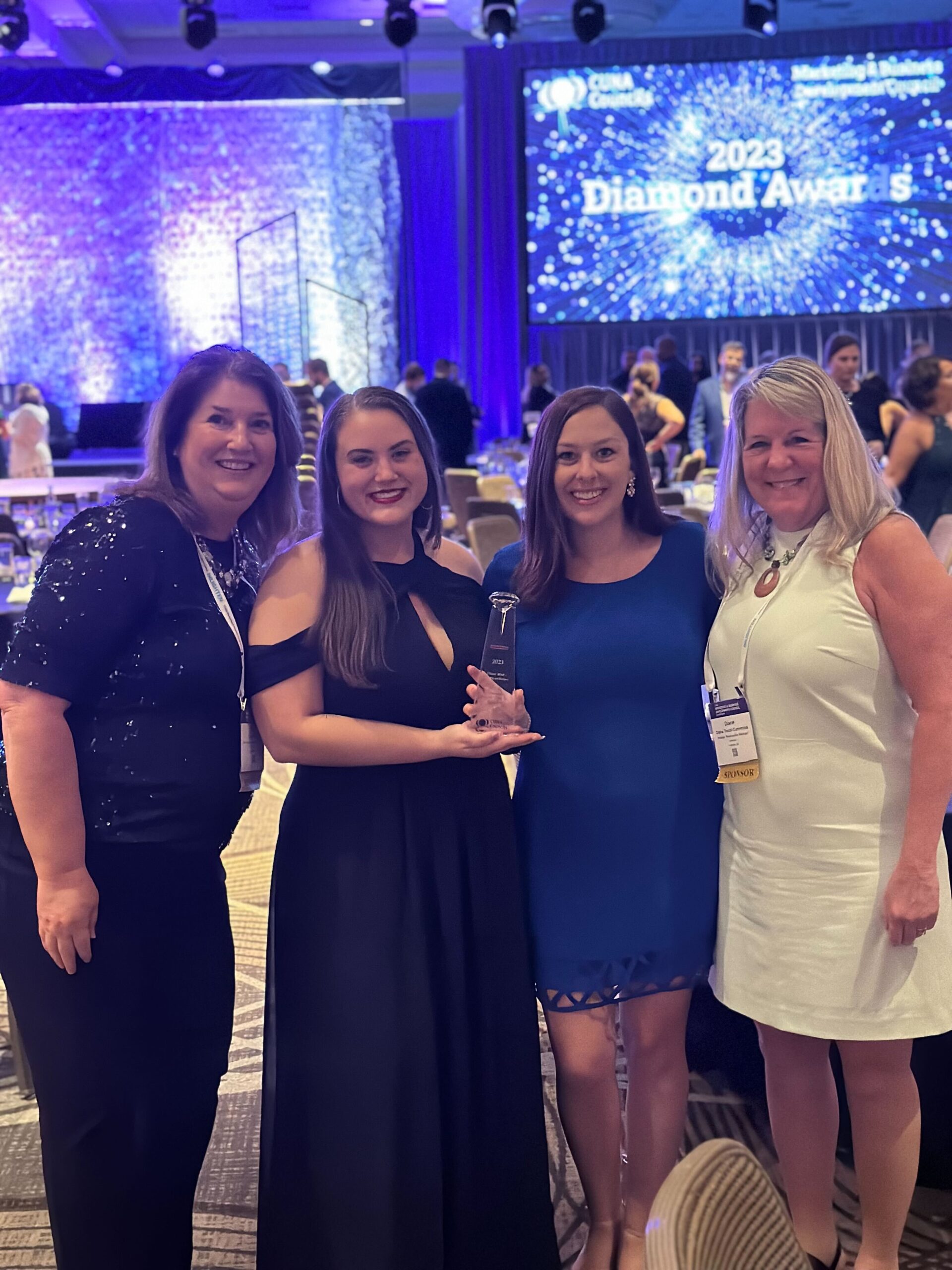 4 women holding a glass trophy, smiling with pride, celebrating their CUNA award victory in a conference hall. 