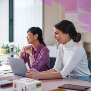 Two women at a conference room table. One is wearing a purple shirt. The other is wearing a white shirt and working on a laptop. Both are smiling.