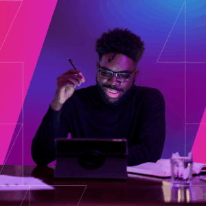 A man wearing glasses and a black sweater smiles while working on a tablet at a desk with papers, a notebook, and a glass of water. The scene is lit with vibrant pink and blue lighting, creating a modern, creative atmosphere.