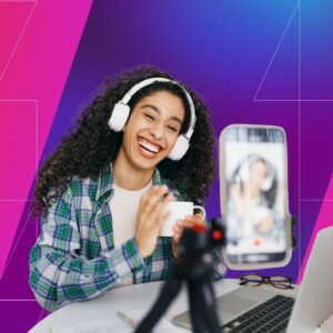 A woman wearing white headphones smiles and gestures toward a smartphone mounted on a tripod while holding a mug, with a laptop and glasses on the table, set against a pink and purple gradient background, suggesting livestreaming or virtual content creation.
