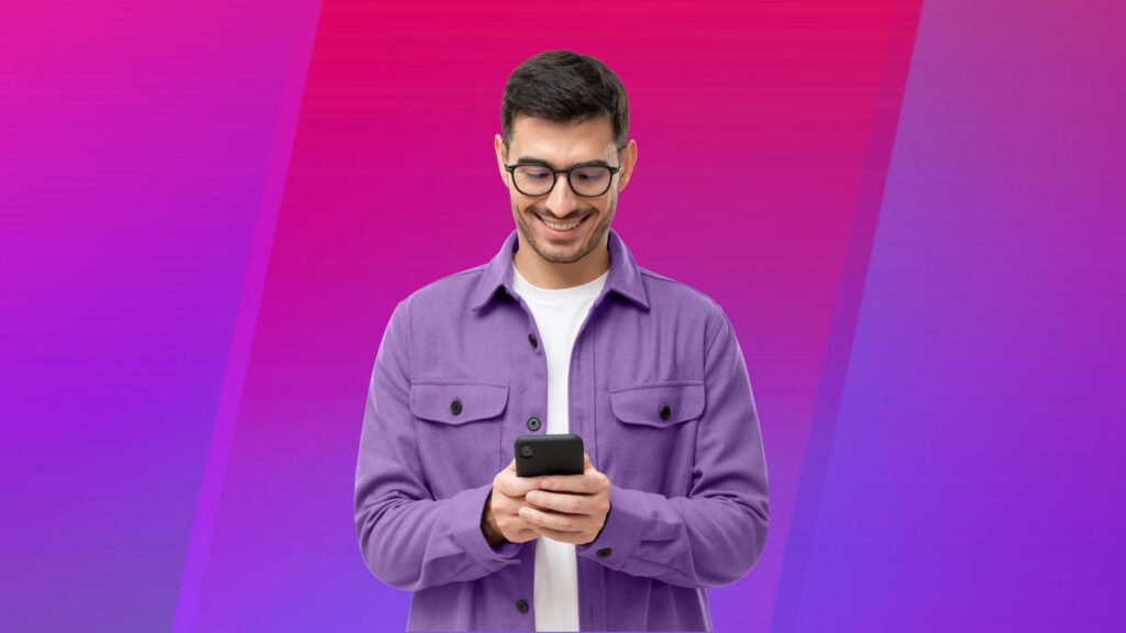 A man wearing glasses and a purple overshirt smiles while looking down at his smartphone, set against a pink and purple gradient background, suggesting mobile engagement or digital communication.