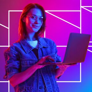 A woman wearing glasses and a denim jacket uses a laptop while illuminated by pink and blue lighting, set against a vibrant geometric background, suggesting digital learning, technology use, or online engagement.