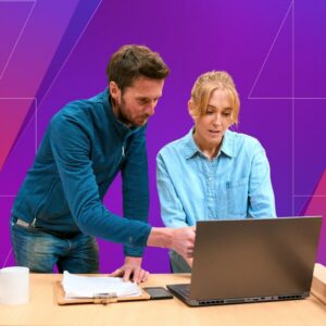 Two coworkers review information on a laptop and printed documents at a table, with one pointing toward the screen, against a purple geometric background.