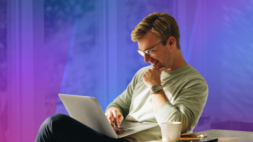 A man wearing glasses smiles while working on a laptop, seated beside a coffee mug and earbuds against a blue and purple gradient background.