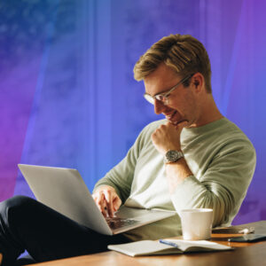 A man wearing glasses smiles while working on a laptop, seated beside a coffee mug and earbuds against a blue and purple gradient background.